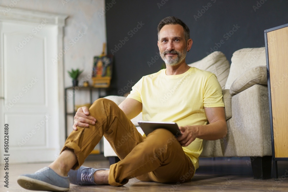 Fototapeta premium portrait of adult man dressed in yellow t-shirt sitting on the floor at home, leaning back on sofa with tablet pc in his hands. Looking at camera and smiling