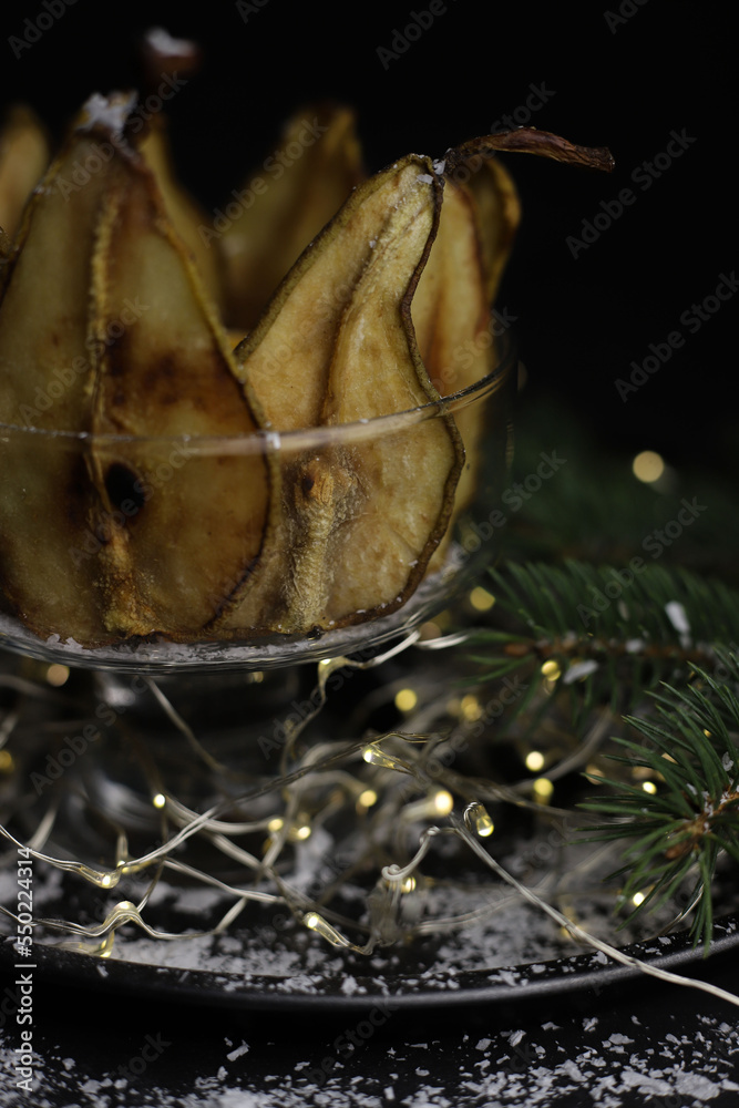 on a dark background and on a black tray stands a transparent glass with dry pears and a branch of a Christmas tree and garlands on top of a little snow. for screensavers, postcards, labels, invitatio