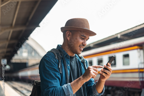 Traveler young asian man using cellphone booking trip at terminal train station. Happy tourist travel by train using smartphone searching location. Male Backpacker arrival at platform railway.