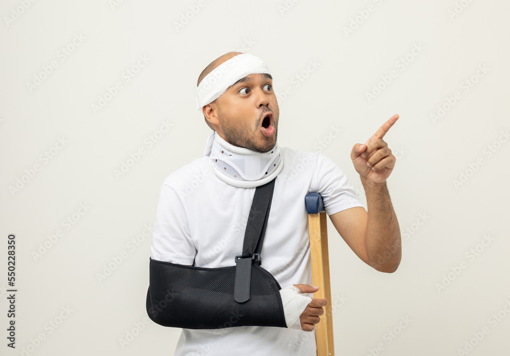 Shocked face man broken arm and leg pointing finger to blank space. Man put on plaster cast splint with walking sticks crutches. Patient wearing sling support arm with neck collar. life insurance Shocked face man broken arm and leg pointing finger to blank space. Man put on plaster cast splint with walking sticks crutches. Patient wearing sling support arm with neck collar. life insurance