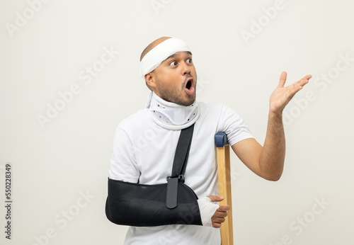 Shocked face man broken arm and leg pointing finger to blank space. Man put on plaster cast splint with walking sticks crutches. Patient wearing sling support arm with neck collar. life insurance