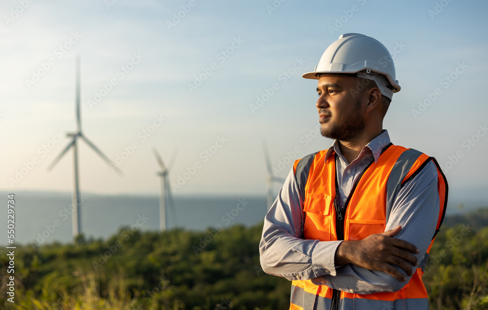 Engineer India man working at windmill farm Generating electricity clean energy. Wind turbine farm generator by alternative green energy. Asian engineer checking control electric power Engineer India man working at windmill farm Generating electricity clean energy. Wind turbine farm generator by alternative green energy. Asian engineer checking control electric power