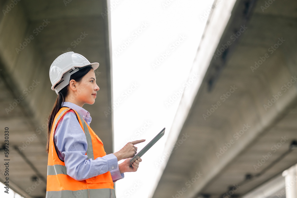Asian engineer worker woman or architect looking construction with tablet wear white safety helmet in construction site. Standing at highway concrete road site. Progress planning of highway bridge. Asian engineer worker woman or architect looking construction with tablet wear white safety helmet in construction site. Standing at highway concrete road site. Progress planning of highway bridge.