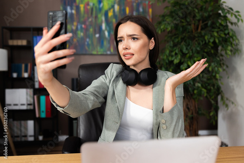 Disappointed worried buisness woman holding cellphone speaking via video link wearing modern earphones and smart suit sitting at desk in comfortable office chair having conferance with colleaguges. photo