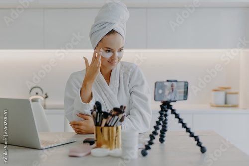 Lovely young female model advertises for internet views cosmetic product cares about skin in under eye area applies hydrogel patches records video on smartphone device for her blog. Vlogging concept photo