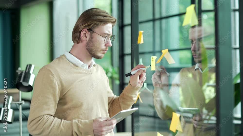 Busy bearded business man employee work with tablet gadget write idea on paper sticker sticky notes on glass wall develops a strategy for startup enterprise financial growth at modern office workplace