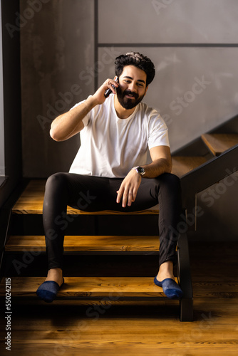 Business man sitting indoors on stairs near flower pots with plants and talking on the phone. Portrait of a business man talking on the phone in a cozy room.