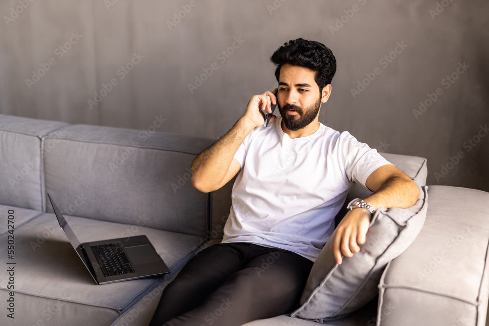 Fototapeta premium Cheerful arab man with long hair and glasses relaxing on couch at home, having phone talk with friends, looking at copy space and smiling, wearing casual outfit, resting at weekend