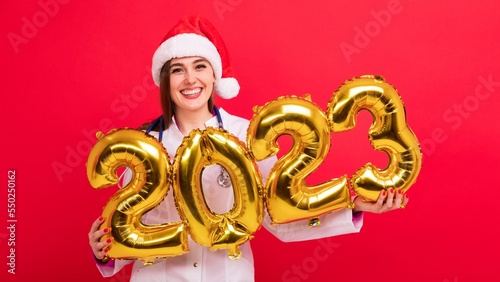 A portrait of a joyful nurse in a white coat and a Santa Claus hat holds the figures of the new year 2023 from balloons on a red background