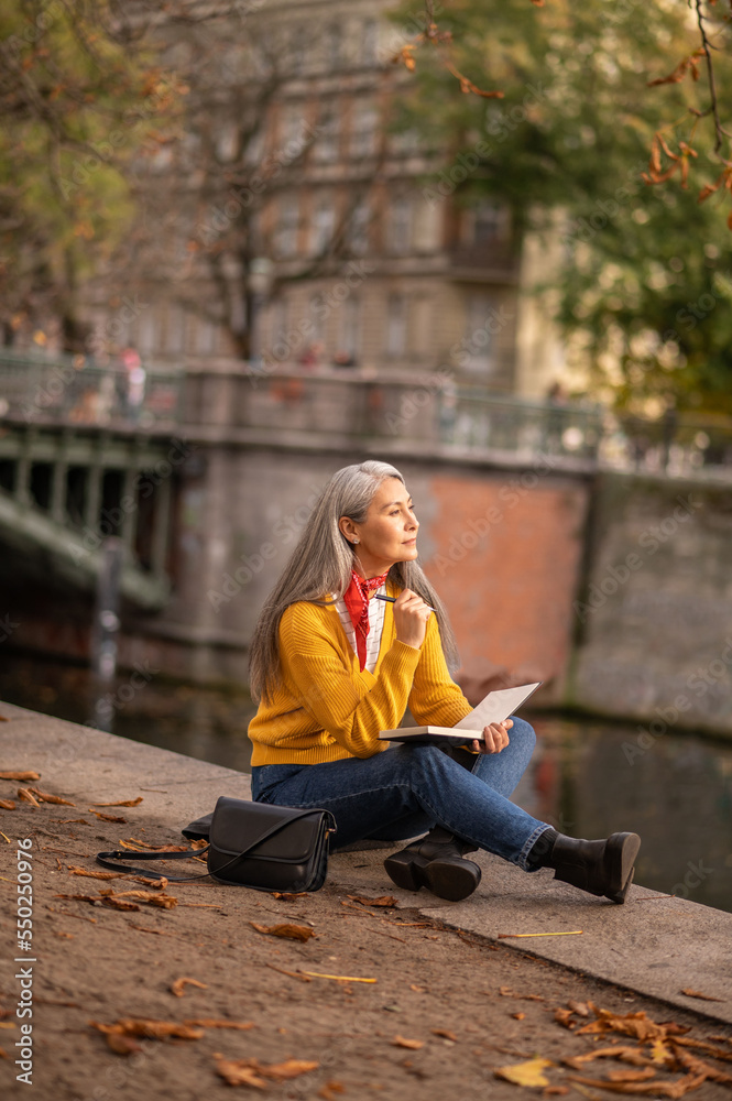 Mature woman sitting on the river bank and drawing in album Mature woman sitting on the river bank and drawing in album