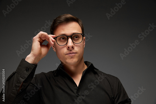 Serious young man  manager in black business classic style shirt putting eyewear on isolated over grey background. Fashion look of businessman. Office style