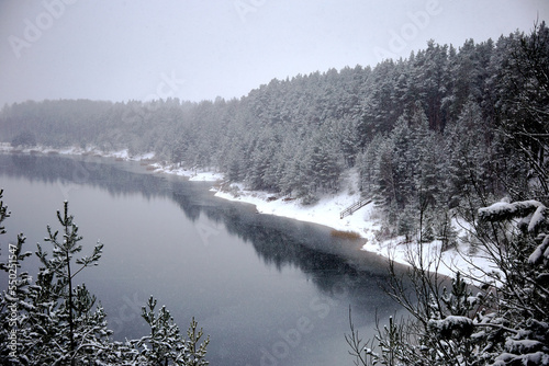 Falling snow flakes above the lake. Winter landscape with forest on the lake shore during the snowfall, selective focus