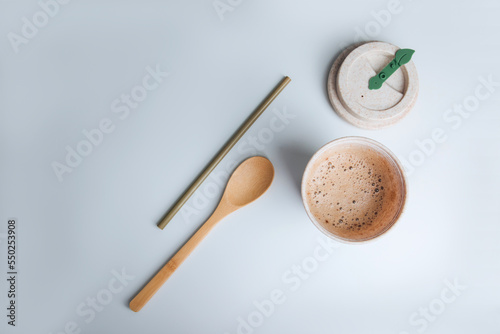 Coffee with foam in an eco glass on a white table with a bamboo straw and wooden spoon. Flat lay