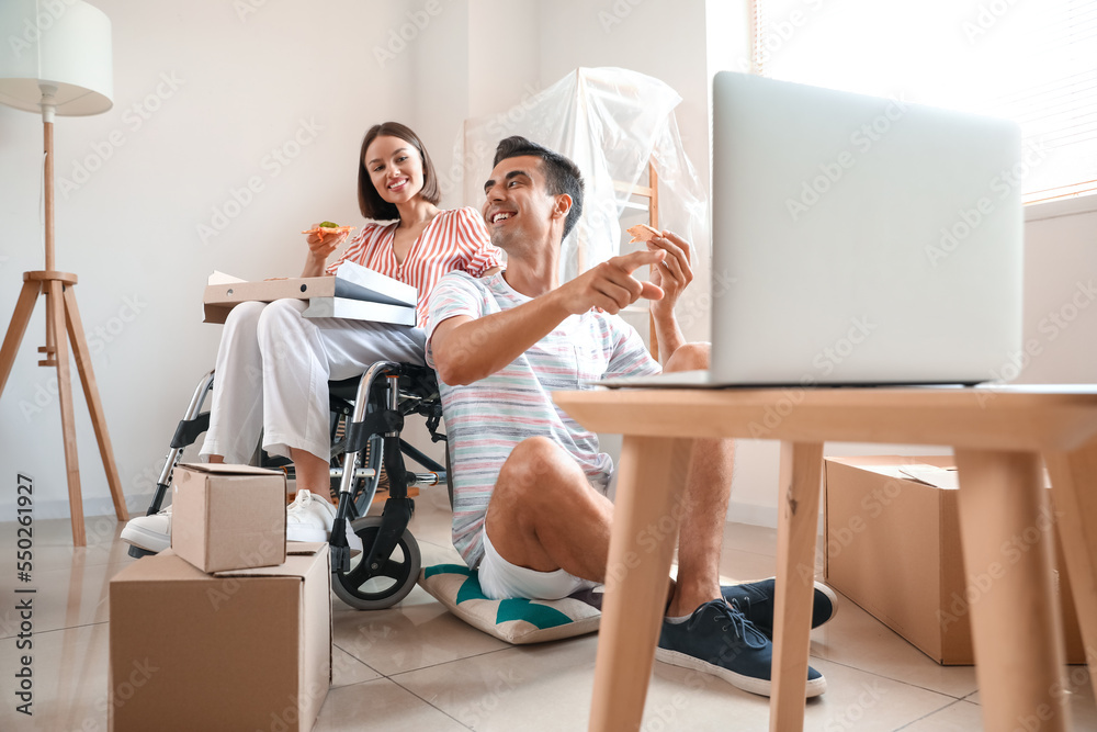 Young man and his wife in wheelchair with pizza watching video in room on moving day
