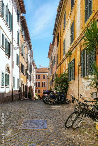A narrow Italian street  with bicycles  motorcycles  and potted plants on the right. Ahead there is a building with yellow stucco.