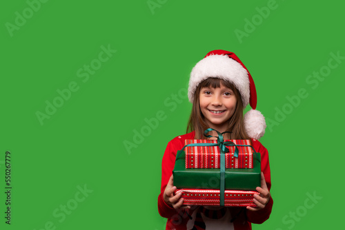 A little smiling girl in a Santa hat is holding a lot of gift boxes. New Year's gifts for Christmas. 2023. Isolated on a green background
