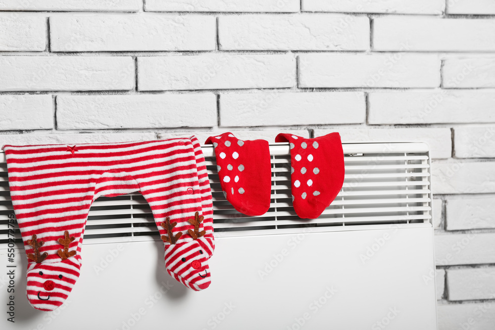 Obraz premium Baby christmas reindeer footed pajamas and red polka dot socks drying on white radiator against brick wall, the festive infant clothing hangs against a textured white brick wall