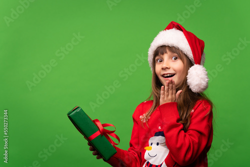 Cute smiling little Santa girl in a hat holds a New Year's gift in her hands and looks happily at the camera. Time to give gifts. Merry Christmas and Happy New Year. Isolated on green background