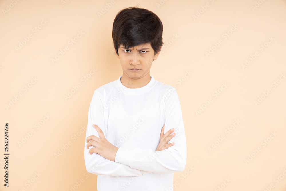 Portrait of angry Indian boy kid wearing white t-shirt with folded arms looking at camera isolated on beige studio background. Asian male child annoyed hands crossed posing , feeling dissatisfied Portrait of angry Indian boy kid wearing white t-shirt with folded arms looking at camera isolated on beige studio background. Asian male child annoyed hands crossed posing , feeling dissatisfied