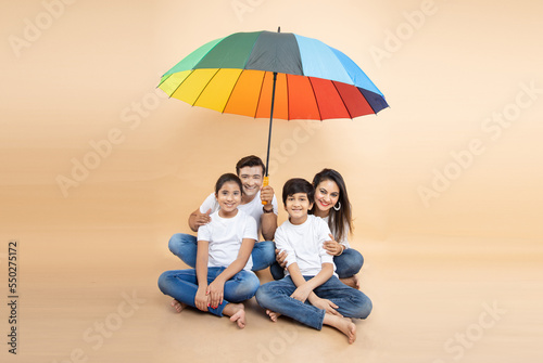 Happy indian family sitting under big multicolor or colorful umbrella isolated on beige background. parents and children. Life and health insurance Safety concept.