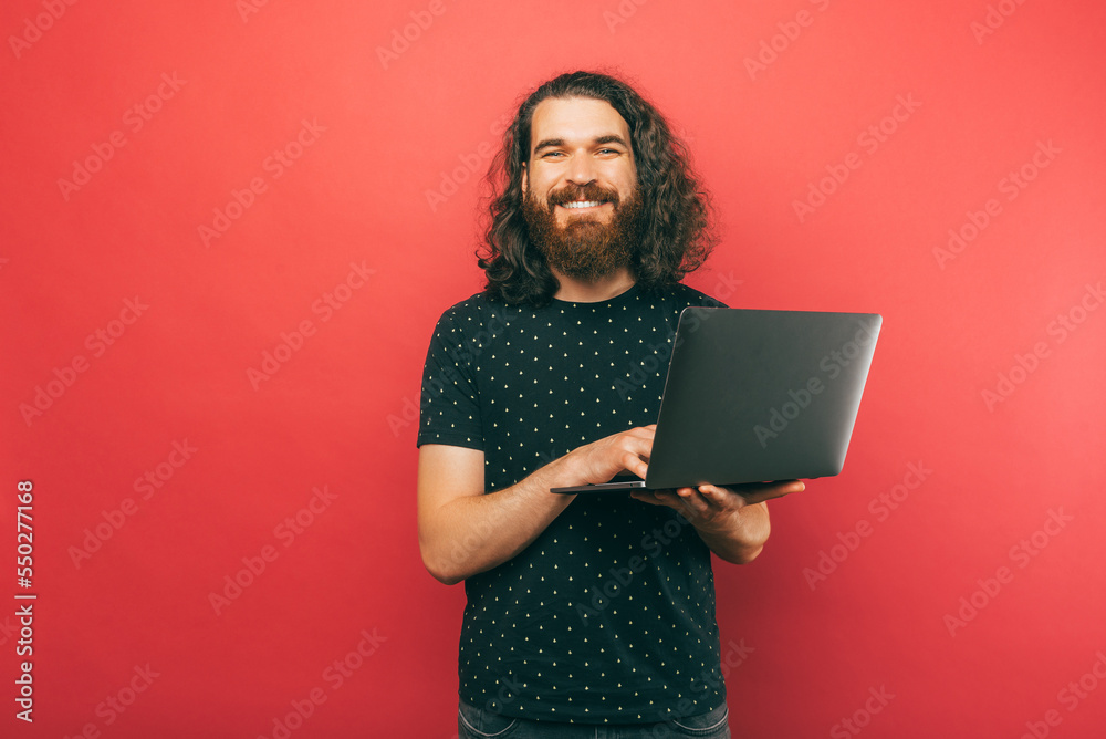 Male bearded hipster is holding a laptop and smiling at the camera in a studio. Male bearded hipster is holding a laptop and smiling at the camera in a studio.