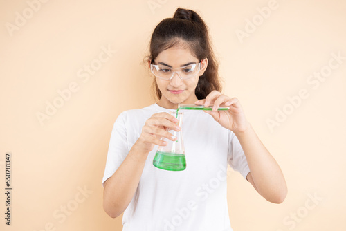 Young indian girl student doing science experiment mixing colorful chemical in a flask isolated over beige background. Education concept.