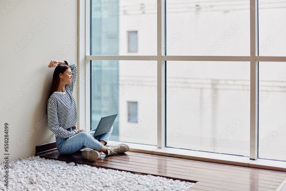 A young woman sits on the floor with her laptop by the window working and studying in the big city