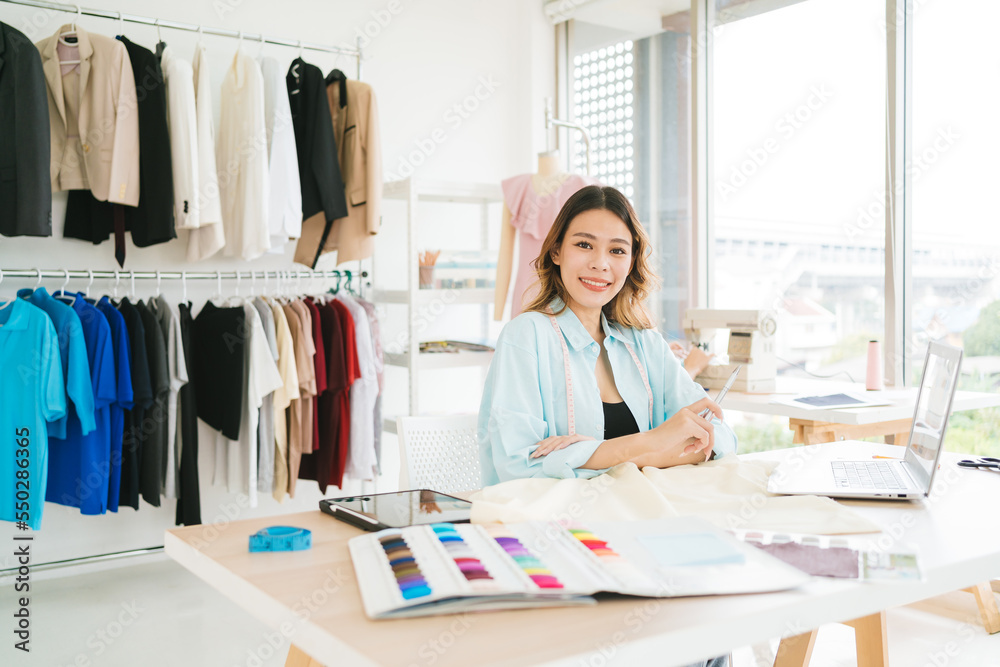 Portrait of a confident Asian tailor woman working on clothes in tailoring atelier. A young attractive female fashion designer smile and look at the camera with a dress-on mannequin in the background. Portrait of a confident Asian tailor woman working on clothes in tailoring atelier. A young attractive female fashion designer smile and look at the camera with a dress-on mannequin in the background.