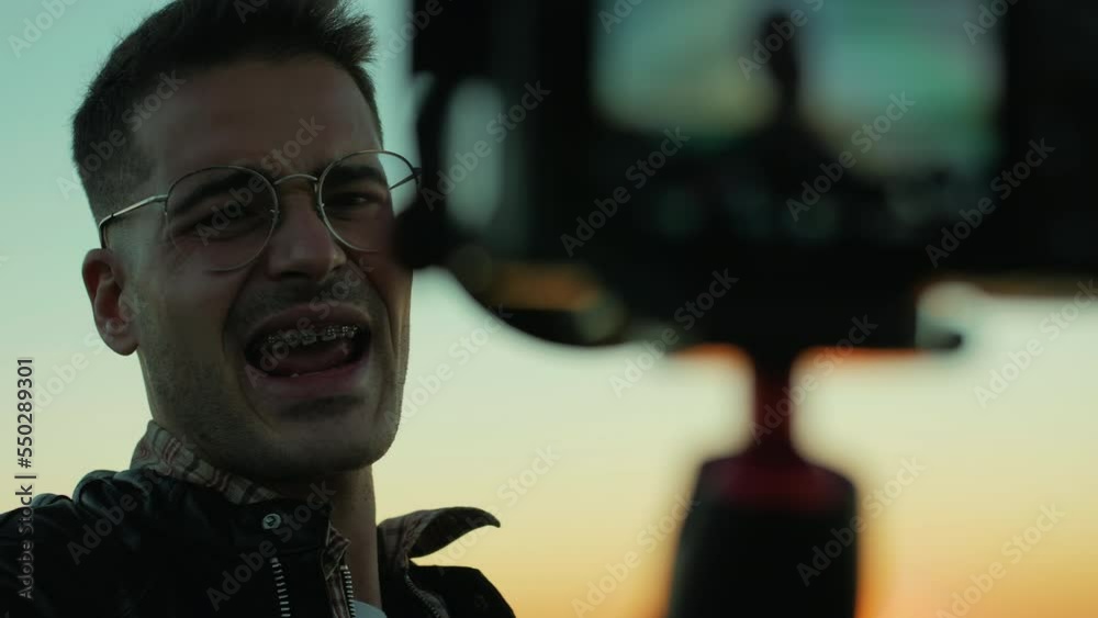 Genuine, handsome generation Z millennial talking to the camera during sunrise. Slow motion of a young, good-looking influencer with dental braces and glasses, vlogging with a camera during sunrise.