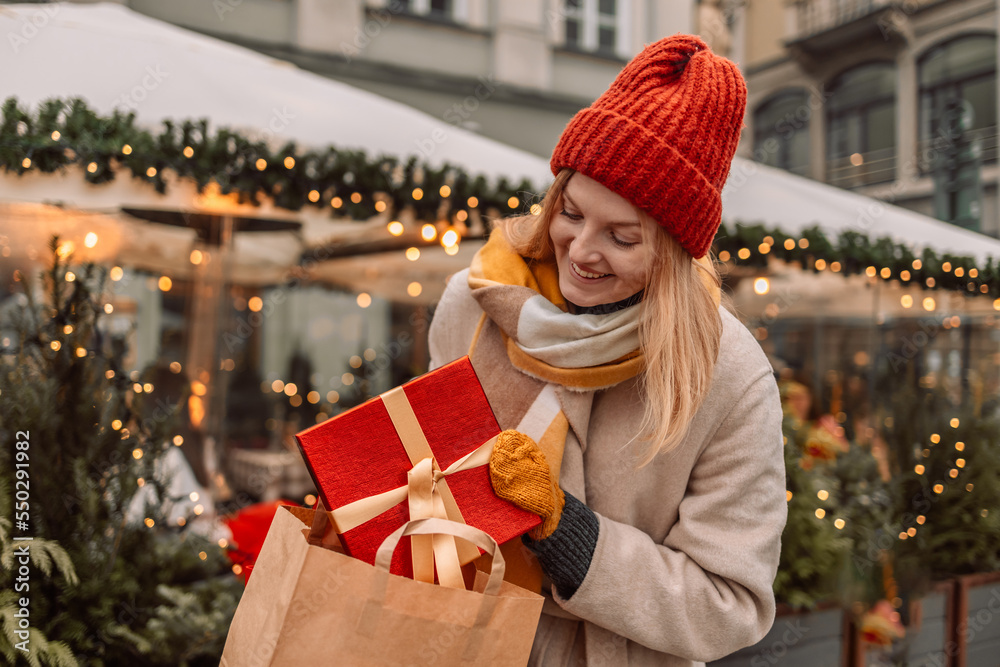 Happy young winter woman with christmas gift. Gift Box Outdoors. Surprise. Cheerful attractive girl shopper with paper bags and presents. Christmas shopping in mall. Winter sales