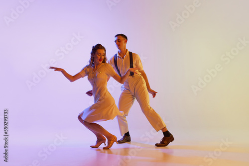 Incendiary dance. Emotional couple of dancers in retro style outfits dancing social dances isolated on gradient lilac color background in neon light. Concept of art, 60s, 70s culture photo