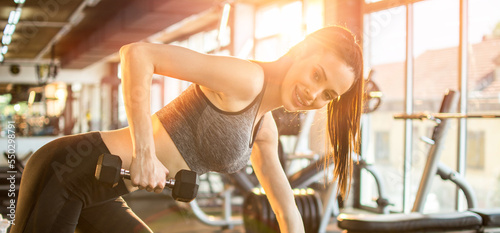 Young fit woman in sportswear working out with weights over fitness bench in gym. Attractive smiling girl working out in front of sunlight in the morning.