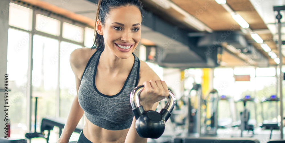 Fototapeta premium Smiling young woman working out with kettle bell in gym. Beautiful fitness girl in sportswear working out with kettle bell in the morning.