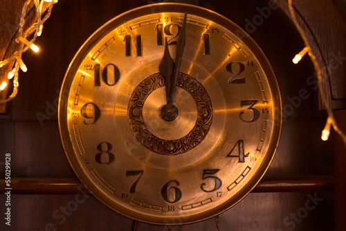 Close-up of the golden dial of an old clock pointing to midnight, illuminated by the warm light of Christmas lights.