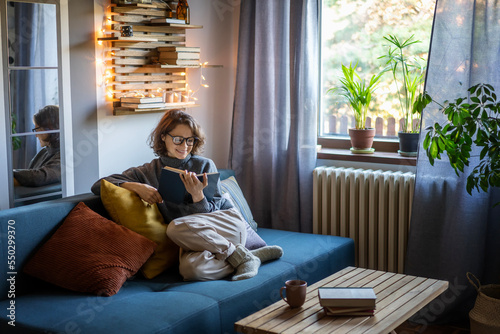 Young smiling cheerful woman in a warm sweater and eyeglasses reading a book while sitting on the couch in the room photo