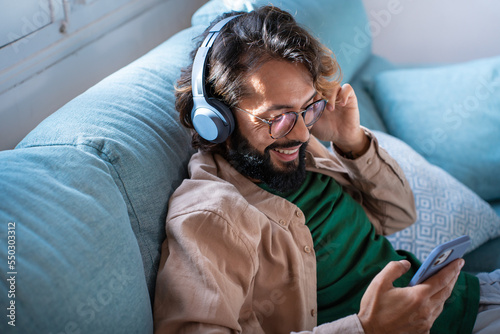 Smiling young man wearing headphones listen to mobile music playing in smartphone app, happy guy relaxing holding using phone enjoy favorite songs relaxing sit on sofa at home. High quality photo photo