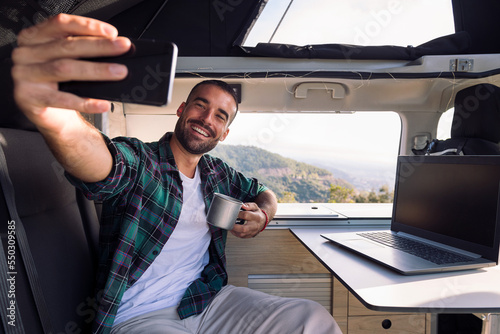 smiling young man taking a selfie inside his camper van using his mobile phone, concept of freedom and digital nomad lifestyle