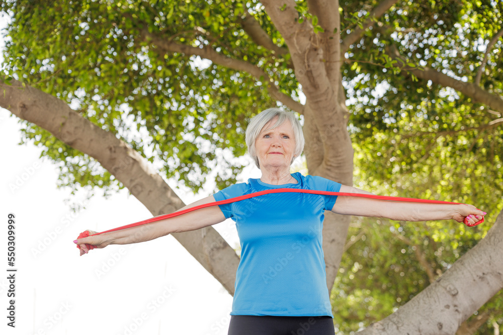 Donna anziana con i capelli grigi e un maglietta sportiva celeste fa esercizio fisico con un elastico rosso  in un parco.
