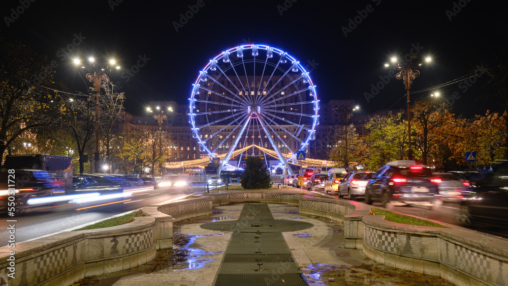 Bucharest ferris wheel for Christmas market, in front of the Parliament building, with cars going by at rush hour. Night, lights, illuminated, symmetry.