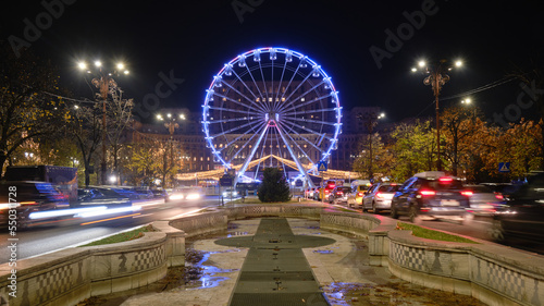 Bucharest ferris wheel for Christmas market, in front of the Parliament building, with cars going by at rush hour. Night, lights, illuminated, symmetry.
