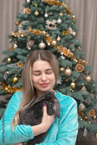 A beautiful Caucasian girl gently holds a plush rabbit in her arms against the backdrop of a Christmas tree. © Fedash