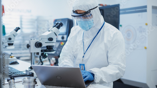 Obraz na płótnie Electronic Manufacturing Factory: Engineer in Sterile Coverall Working on Laptop Computer, Examining a Circuit Board with Microchips Through a Microscope and Testing New Electrical Equipment