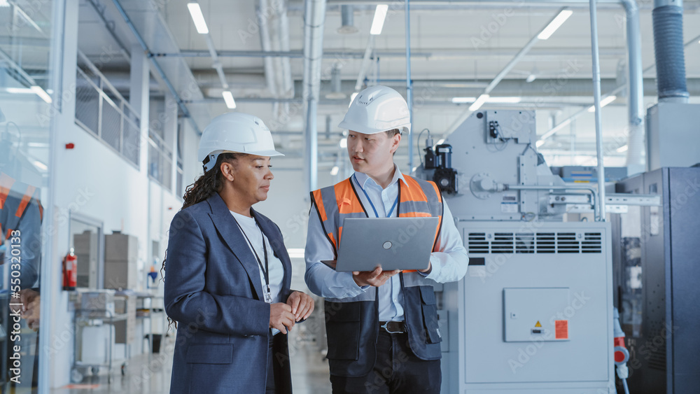 Two Professional Heavy Industry Engineers Walking at a Factory in Safety Uniform and Hard Hats, Discussing Work on Laptop Computer. Asian Specialist and African American Technician at Work. Two Professional Heavy Industry Engineers Walking at a Factory in Safety Uniform and Hard Hats, Discussing Work on Laptop Computer. Asian Specialist and African American Technician at Work.