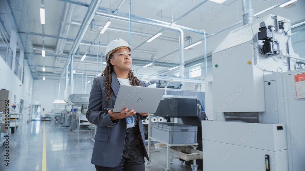 Portrait of a Black Female Engineer in Hard Hat Standing and Using Laptop Computer at Electronic Manufacturing Factory. Technician Working on Daily Tasks and Research and Development Data. Portrait of a Black Female Engineer in Hard Hat Standing and Using Laptop Computer at Electronic Manufacturing Factory. Technician Working on Daily Tasks and Research and Development Data.
