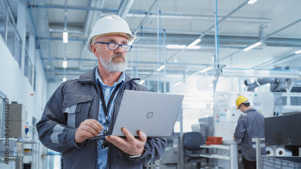 Portrait of a Bearded Middle Aged General Manager Standing in a Factory Facility, Wearing Work Jacket and a White Hard Hat. Heavy Industry Specialist Working on Laptop Computer. Portrait of a Bearded Middle Aged General Manager Standing in a Factory Facility, Wearing Work Jacket and a White Hard Hat. Heavy Industry Specialist Working on Laptop Computer.