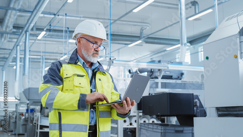 Portrait of a Bearded Middle Aged Engineer Standing in a Factory Facility, Wearing a High Visibility Jacket and a White Hard Hat. Heavy Industry Specialist Working on Laptop Computer.