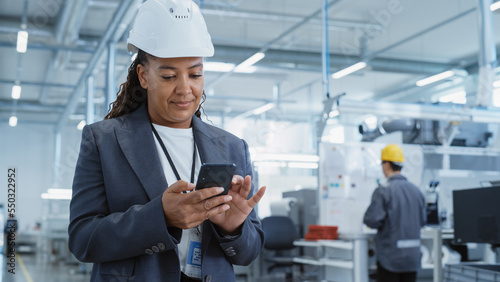 Portrait of a Black Female Engineer in Hard Hat Standing and Using a Smartphone at Electronics Manufacturing Factory. Technician is Writing a Message and Checking Her Schedule.