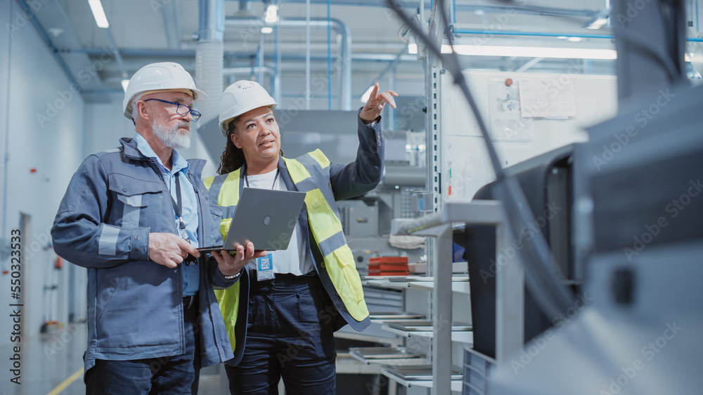 Portrait of Two Heavy Industry Employees in Hard Hats at Factory. Checking and Discussing Industrial Facility, Using Laptop Computer. African American Engineer and Middle Aged Technician at Work. Portrait of Two Heavy Industry Employees in Hard Hats at Factory. Checking and Discussing Industrial Facility, Using Laptop Computer. African American Engineer and Middle Aged Technician at Work.