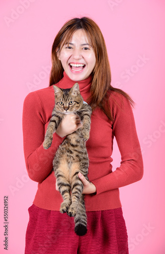 A young asian pretty cute woman in a red sweater holds a cat in her hands like a baby isolated on a bright color pink background. Asia girl with a kitten. Fiendship of pet and owner. photo