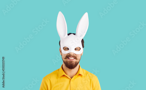 Portrait of cheerful and funny bearded man in rabbit carnival mask isolated on turquoise background. Close up of young man in white rabbit mask with long ears smiling looking at camera. Banner.